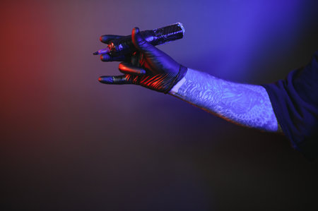 Closeup of tattooer master's hand in black glove holding machine for making tattoo art on body isolated on dark background in neon. Concept of art, job, hobby, professional equipment. Flyer for adの写真素材