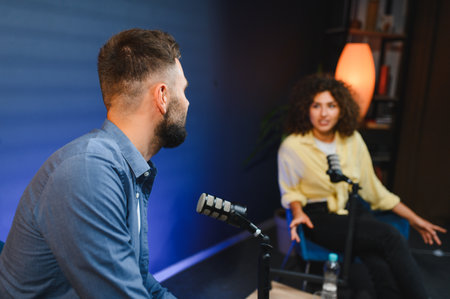 Two podcasters, a man and a woman, are recording a podcast in a professional studio, using...