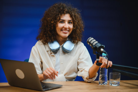 Happy female podcaster speaking into microphone while recording audio podcast in studio with blue...