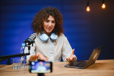 Young woman recording a vlog using a professional microphone, headphones, and laptop in a studio...