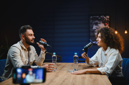 Two content creators, a man and a woman, are recording a podcast in a professional studio, using microphones and a smartphone for live streamingの写真素材