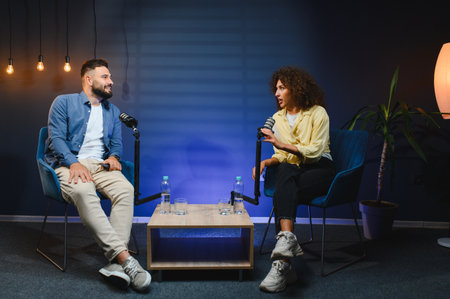 Two podcasters, a man and a woman, are recording a podcast in a professional studio, engaging in...