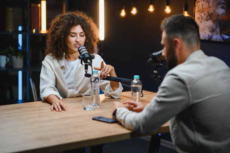 Two podcasters, a man and a woman, are recording audio content in a professional studio, using...