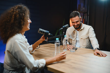 Two happy colleagues are recording a podcast together, laughing and talking into microphones in a...