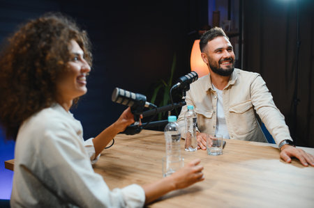 Two smiling podcasters recording audio content in professional studio, using microphone and...