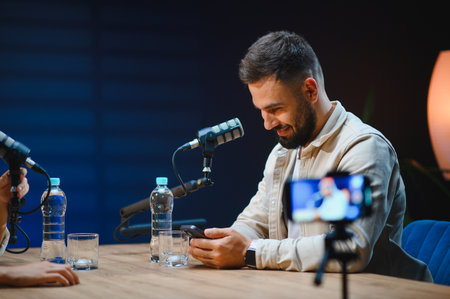 Two content creators recording podcast episode and streaming live video using smartphone and professional microphone in modern studioの写真素材