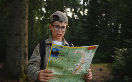 Young boy scout wearing glasses and carrying backpack reading map in forest, trying to find way back...