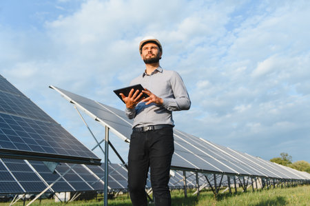 Engineer wearing a hardhat and holding a tablet, inspecting a solar panel field for renewable energy...