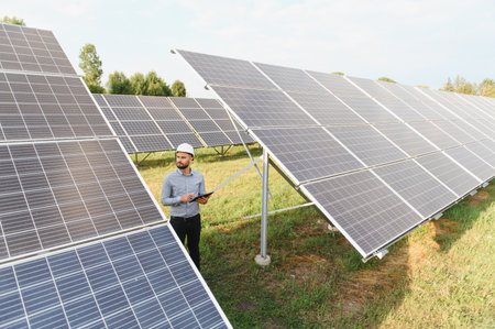 Engineer wearing a hard hat and holding a tablet, inspecting a field of solar panels for sustainable...