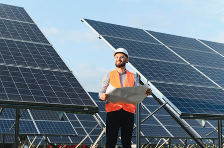 Male engineer working at a solar power plant, holding a blueprint, analyzing renewable energy...