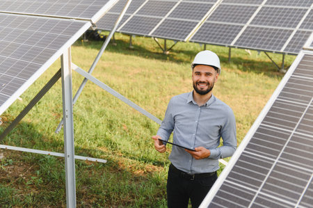 Smiling engineer wearing a hard hat, checking the efficiency of solar energy panels at a modern...