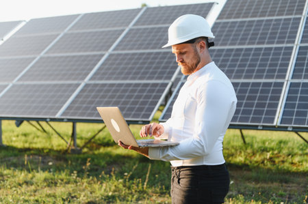 Engineer wearing hard hat checking solar panel performance and analyzing data on a laptop in a solar...