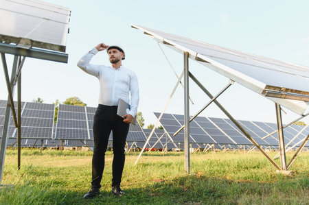 Man in a hard hat holding a laptop, standing in a large solar panel field and looking at the...
