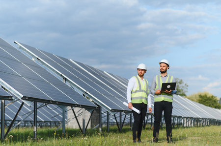 Two engineers in safety gear review solar panel operations with a laptop at a renewable energy plant...