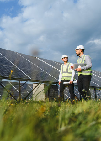 Two engineers in safety gear inspect large solar panels, discussing renewable energy solutions under...