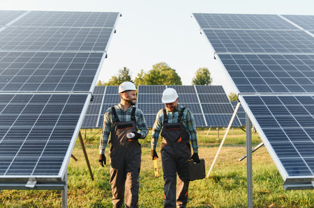 Engineers in hard hats and overalls walking and talking through a solar panel field, installing new...