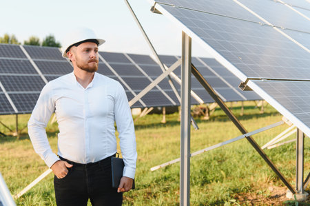 Male engineer in hard hat inspecting rows of solar panels at a large photovoltaic solar farm,...
