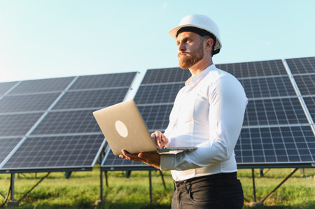 Engineer wearing a hard hat checking renewable energy system performance on a laptop at a solar power plantの写真素材