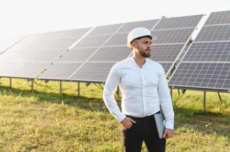 Engineer man in a hard hat standing, holding a laptop, observing a large solar panel field under a...