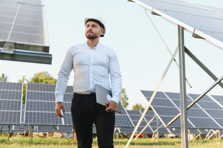 Man in hard hat holding laptop, overseeing solar farm operations, integrating technology for...