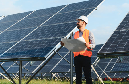 Young male engineer wearing a hard hat and safety vest, holding blueprints while inspecting solar...