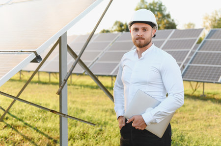 Man engineer wearing hard hat standing in a solar farm, holding a laptop. Representing...