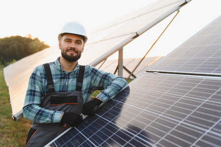 Male technician wearing a hard hat and overalls, confidently smiling while inspecting a solar panel...