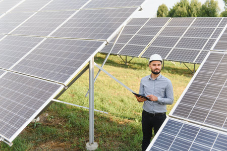 Engineer wearing hard hat holding a tablet and inspecting solar panel array, focusing on renewable...