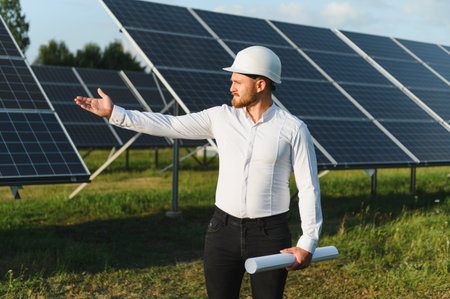 Engineer wearing a hard hat, holding blueprints, and pointing at solar panels, representing...