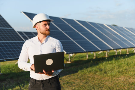 Engineer wearing a hard hat and holding a laptop, standing in front of solar panels at a power plant...