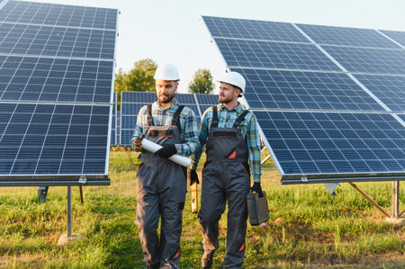 Two skilled engineers in hard hats and overalls discussing plans while walking through a large solar...