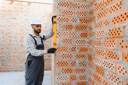 Bricklayer checking vertical alignment of new brick wall with a spirit level, ensuring quality...