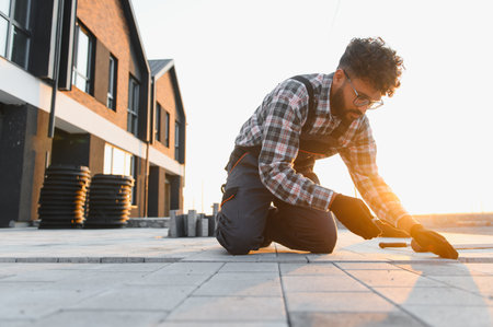 Construction worker kneeling, carefully laying paving stones on the ground at a building site during sunsetの写真素材