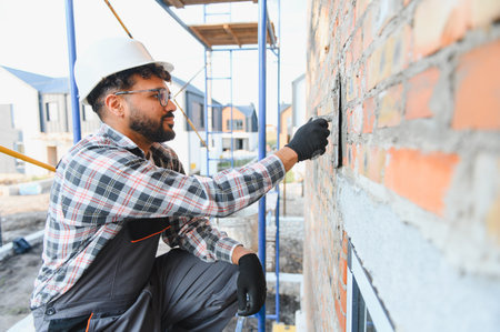 Young male worker wearing hard hat and workwear, applying mortar to a brick facade on a construction...