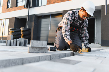 Worker in hard hat checking the level of new paving stones during construction project, ensuring precise outdoor installationの写真素材