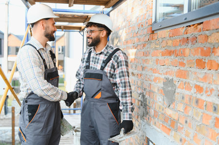 Two smiling male builders in hard hats and overalls performing a handshake at a construction site,...