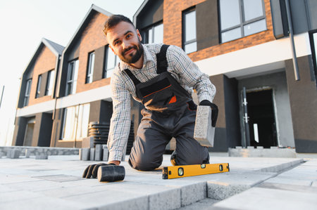 Young construction worker installing patio paving with a level and hammer in front of new modern...