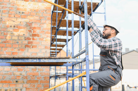 Worker in hard hat and overalls climbing metal scaffolding near a brick wall, focusing on...