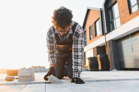 Man installer in overalls and plaid shirt kneeling to lay paving stones on a residential driveway under warm sunset lightの写真素材