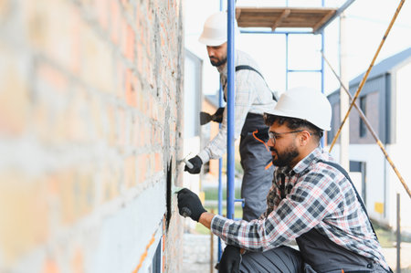 Construction workers wearing hard hats and safety gear plastering a brick wall on scaffolding at a...