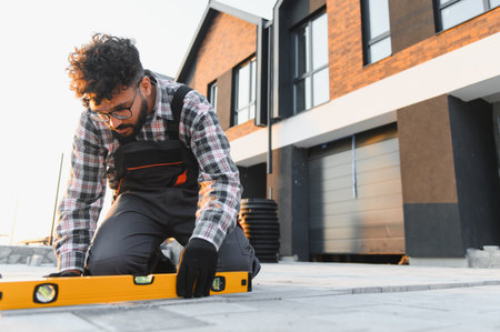 Man kneeling, carefully checking the level of paving stones. Building a new driveway for a modern...