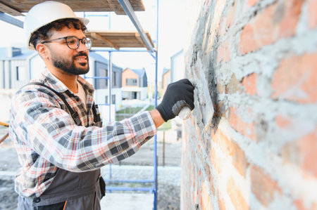 Construction worker in hard hat applying mortar with trowel to brick wall, building new residential homes at job siteの写真素材