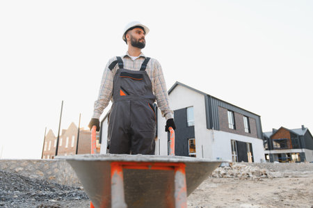 Young man in hard hat and overalls moving materials with a wheelbarrow on a new house construction siteの写真素材