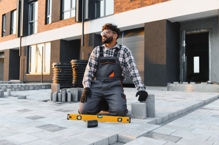 Young male tradesman laying concrete paving blocks by a new townhouse, using a level tool to ensure...