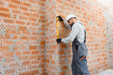 Professional construction worker wearing hard hat and overalls, using a spirit level on a brick...