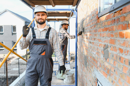 Construction worker in hard hat and overalls making an okay sign, with another builder plastering a...