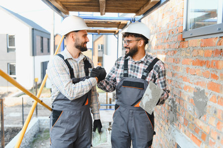 Construction workers wearing hard hats and overalls on a building site, fist bumping to celebrate...