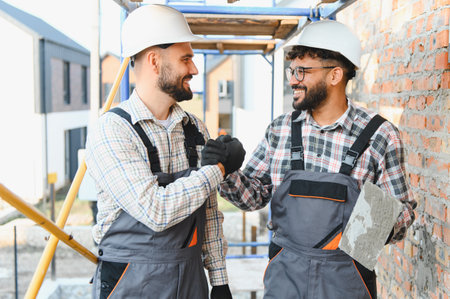 Two diverse builders wearing hard hats, shaking hands, symbolizing successful teamwork and collaboration at a construction siteの写真素材