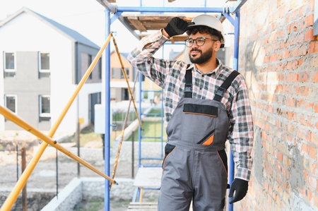 Young male builder on construction site wearing hard hat and overalls, overseeing new residential...
