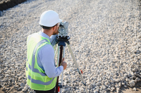 Engineer wearing safety vest and hard hat using a total station for land measurement on a gravel...